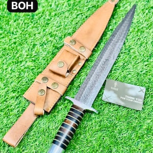 Può includere: Un pugnale con una lama a motivi e un manico con strisce marroni scure e chiare alternate. Il pugnale è accompagnato da un fodero in pelle marrone chiaro con rivetti color ottone. Il testo "BOH" si trova nell'angolo in alto a sinistra.