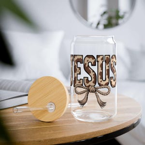 Op de afbeelding: Een doorzichtige glazen beker in de vorm van een blikje met het woord "JESUS" in een bruin en beige dierenprint. Onder het woord staat een touwstrik. De beker heeft een bamboe deksel en een doorzichtig rietje.