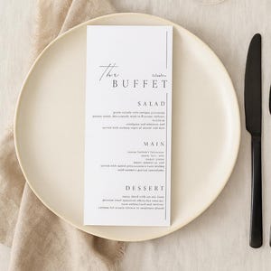 Könnte beinhalten: Eine minimalistische Menükarte mit dem Text "The Buffet" und einer Liste von Gerichten, platziert auf einem weißen Teller mit einem schwarzen Messer. Ein Weinglas und eine beige Serviette sind ebenfalls sichtbar.