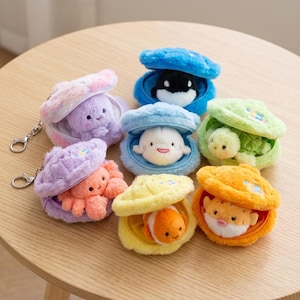 May include: A collection of six plush sea creature keychains, each nestled inside a soft, fuzzy clam shell. The creatures include an octopus, orca, beluga whale, sea turtle, clownfish, and pufferfish, all featuring embroidered facial details.