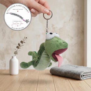 Pode incluir: Um porta-chaves de peluche em forma de sapo verde com olhos brancos grandes e uma língua cor-de-rosa de fora. O sapo está preso a um anel de metal prateado por uma corrente curta. Um gráfico circular exibe texto e um pendente de missangas.