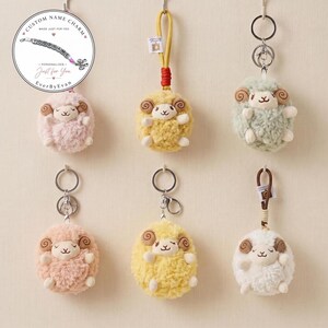 May include: Six plush sheep keychains in various colours including pink, yellow, green, peach, and white. Each sheep features curled brown horns, a smiling face, and a metal keyring attachment. A circular logo in the top left reads Custom Name Charm.