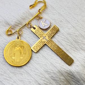 Puede incluir: Alfiler de seguridad dorado con amuletos religiosos. Incluye un colgante de cruz con texto grabado, una medalla redonda y un amuleto con la letra "K". Accesorio decorativo.