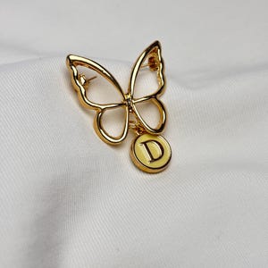 Op de afbeelding: Goudkleurige vlinderbroche met opengewerkte vleugels. Een kleine, ronde bedel met een gele emaille achtergrond en de letter "D" hangt onder de vlinder. De broche is geplaatst tegen een witte, getextureerde achtergrond.