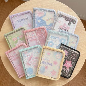 May include: A collection of ten small, rectangular, ring-bound notebooks featuring clear plastic covers decorated with white star patterns. Each notebook contains colorful inserts with various star-themed graphics and words like SHINE, LUCKY, SOFT, ONE LOVE, and BLING.