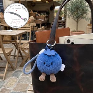 Könnte beinhalten: Ein plüschiger Taschenanhänger in Blaubeerform mit einem flauschigen blauen Körper, einem kleinen grünen Blatt als Oberteil und zwei braunen Beinen. Der Anhänger hat ein einfaches gesticktes Gesicht und ist an einer dunkelbraunen Ledertasche befestigt.