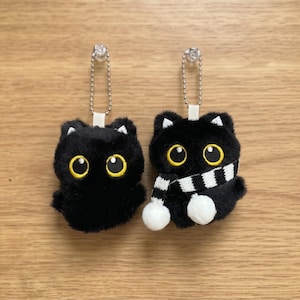 May include: Two plush black cat keyrings with yellow eyes and white accents. One cat has a black and white striped scarf. Each keyring has a silver chain and a white fabric loop for attachment.