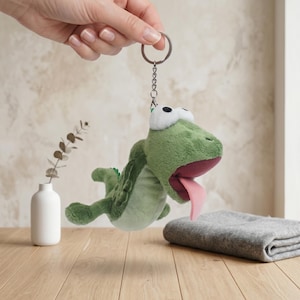 Puede incluir: Un llavero de peluche de cocodrilo verde con lengua rosa y ojos blancos. El cocodrilo está suspendido de una cadena y un llavero plateados. El peluche está sostenido por una mano, con un fondo neutro.