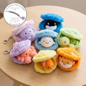 May include: A collection of plush sea animal keychains, each nestled inside a soft, fuzzy, openable shell. The set includes a purple octopus, a blue orca, a white beluga, a green turtle, an orange clownfish, and a yellow pufferfish.