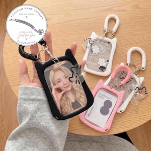 Könnte beinhalten: Eine Sammlung von katzenförmigen Kartenhaltern in Schwarz, Weiß und Pink, jeweils mit einer spiralförmigen Handschlaufe, einem Metallschlüsselring und einem kleinen Katzenanhänger. Der schwarze Halter zeigt ein Foto, die anderen Karten.