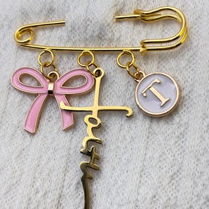 Op de afbeelding: Een goudkleurige veiligheidsspeld met drie bedels. Eén bedel is een roze strik, een andere is het woord "Faith" in een cursief lettertype en de derde is een witte cirkel met de letter "T" in het midden. De speld ligt op een wit, getextureerd oppervlak.