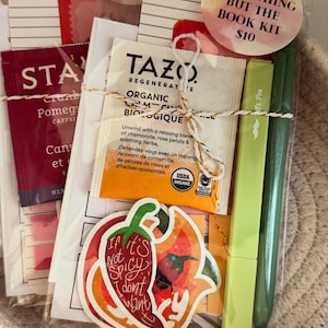 May include: A clear cellophane-wrapped kit containing a Tazo tea bag, two green pens, a sticker with a chili pepper design, and reading journal cards. The kit is tied with gold twine and includes a round sticker that says "Spicy Everything But The Book Kit".