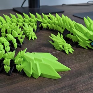 May include: A segmented, articulated sculpture of a stylized dragon in bright green and black. The dragon's body is composed of numerous geometric segments, with a head featuring a blue eye. The sculpture is displayed on a wooden surface.