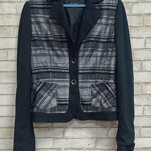 Puede incluir: Un blazer a cuadros negros y grises con mangas negras y cuello negro. El blazer tiene dos bolsillos delanteros y dos botones. La marca "Tuzzi" es visible en la etiqueta.