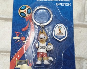 Pode incluir: Um chaveiro com o mascote Zabivaka da Copa do Mundo FIFA 2018 na Rússia, em uniforme vermelho e branco, segurando uma bola de futebol. A embalagem azul inclui o texto "2018 FIFA WORLD CUP RUSSIA" e o logotipo oficial.