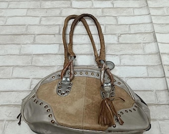 May include: Silver and tan handbag with a rounded shape. The bag features a central tan panel, silver accents, and decorative studs. It has two brown handles and a tassel detail.