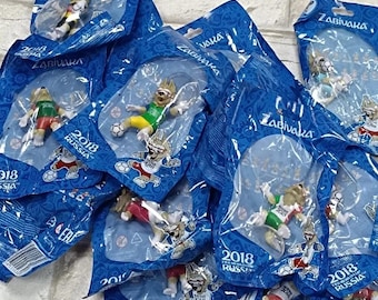 Pode incluir: Uma coleção de figuras ZABIVAKA em embalagens azuis. Cada figura é um lobo de desenho animado vestindo roupas esportivas, representando a Copa do Mundo FIFA 2018 na Rússia. A embalagem inclui o texto "2018 RUSSIA".