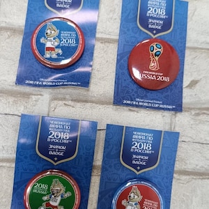 May include: Four circular 2018 FIFA World Cup Russia pin-back badges on blue cardboard backing cards. The badges feature the tournament mascot Zabivaka, the official trophy, and the text 2018 FIFA World Cup Russia in English and Russian.