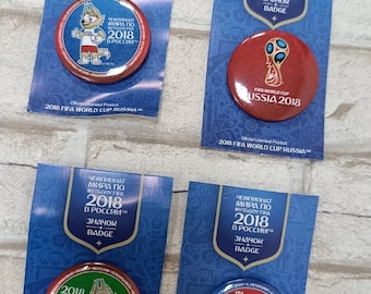 Pode incluir: Quatro crachás redondos 2018 FIFA World Cup Russia em cartões de suporte azuis. Os crachás apresentam a mascote Zabivaka, o troféu oficial e o texto 2018 FIFA World Cup Russia em inglês e russo em cada uma das embalagens.