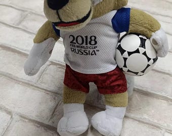 Pode incluir: Um brinquedo de pelúcia de Zabivaka, o mascote oficial da Copa do Mundo FIFA de 2018 na Rússia. O personagem semelhante a um lobo está vestindo uma camisa branca com o logotipo do evento, shorts vermelhos e segurando uma bola de futebol. Possui óculos com bordas laranja.
