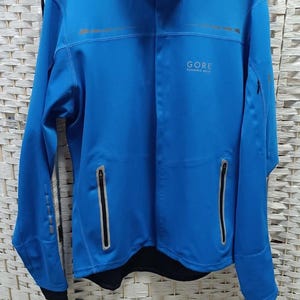 Könnte beinhalten: Leuchtend blaue GORE-Laufjacke mit hohem Kragen und zwei Reißverschlusstaschen vorne. Die Jacke hat schwarze Bündchen und einen reflektierenden Streifen. Das GORE-Logo ist auf Brust und Kragen sichtbar. Sportbekleidung.