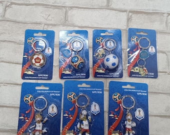 Pode incluir: Uma coleção de chaveiros em cartões azuis. Os chaveiros apresentam bolas de futebol, troféus e uma figura de mascote. Os cartões têm o texto "БРЕЛОК" e "2018 FIFA WORLD CUP RUSSIA". Os chaveiros da mascote têm o texto "ZABIVAKA".