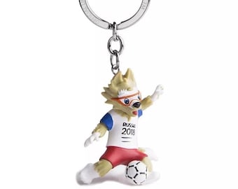 Pode incluir: Um porta-chaves com Zabivaka, a mascote oficial da Copa do Mundo FIFA 2018. A personagem, com forma de lobo, é retratada numa pose de futebol, vestindo uma camisa branca com "RUSSIA 2018" estampada, calções vermelhos e detalhes azuis e brancos.