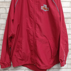 Puede incluir: Una chaqueta roja con capucha y cremallera que presenta el logotipo de Royal Mail en el pecho izquierdo. La prenda incluye una cremallera frontal completa, mangas largas con puños elásticos y detalles de ribetes reflectantes plateados.