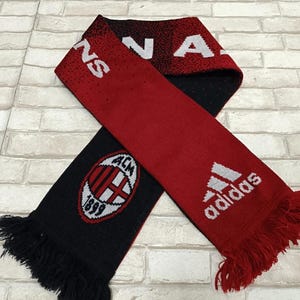 Puede incluir: Bufanda roja y negra con el texto "AC MILAN" y el logo de Adidas. La bufanda tiene un borde con flecos y presenta el emblema del equipo. La bufanda está hecha de un material de punto.