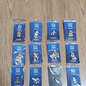 May include: A collection of 2018 FIFA World Cup pin badges, each sealed in a clear plastic bag. The badges feature the official mascot and the tournament logo on blue backing cards. The pins are in various designs and poses.