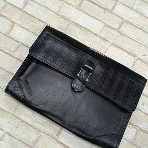 May include: Black leather briefcase with a textured flap and a silver buckle. The rectangular briefcase has a smooth body and a woven pattern on the flap. The corners feature a diamond pattern. The briefcase is made of leather.