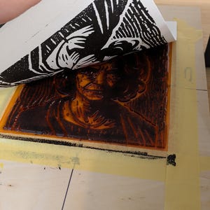 May include: A hand lifts a white paper to reveal a black and white print of a person's face. The print is on a wooden block with a brown ink layer. The block is taped to a wooden surface, showcasing the printmaking process.