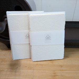 Könnte beinhalten: Zwei Stapel cremefarbenes, strukturiertes Papier mit Büttenrand, jeweils zusammengehalten durch eine weiße Papierbanderole mit einem kleinen Fleur-de-Lis-Logo und dem Text Everbound Press. Das Papier hat eine sichtbare Körnung.