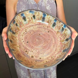 Könnte beinhalten: Eine breite, flache Keramikschale mit unregelmäßigem, organischem Rand. Das Innere zeigt eine strukturierte Glasur mit einem zentralen Wirbel in Altrosa und Beige, der in einen Rand mit strahlenförmigen blauen und dunkelgrünen Tropfen übergeht.