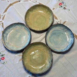 Puede incluir: Un juego de cuatro cuencos de cerámica redondos con bordes irregulares y ondulados. Dos cuencos presentan un esmalte verde salvia claro, mientras que los otros dos muestran un acabado moteado en azul y crema. Cada cuenco tiene un borde de arcilla.