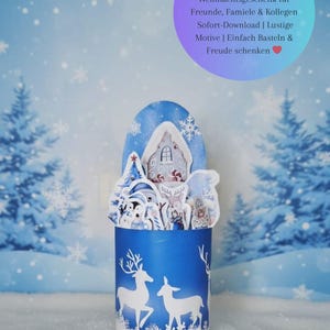 May include: A blue Christmas-themed gift box with a winter scene. The box features white deer silhouettes and the text "Frohe Weihnachten." Inside, there's a miniature winter village with a house, trees, and figures. Text on the image says "DIY Duplo-Banderolen - Perfektes Weihnachtsgeschenk".