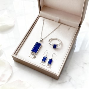 May include: A silver jewellery set comprising a necklace, ring, and earrings, displayed in a gift box. The set features a rectangular, deep blue gemstone pendant on the necklace and earrings, and a square gemstone on the ring. The earrings and necklace have small, round silver accents.