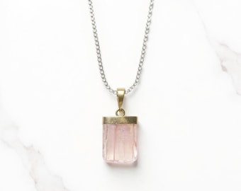May include: A silver chain necklace with a rectangular, light pink pendant. The pendant is encased in a gold-colored metal setting. The necklace is displayed on a white marble surface.