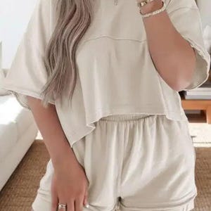 May include: A beige two-piece lounge set. The set includes a short-sleeved, crew neck top and matching shorts. The fabric appears to be a soft, comfortable material. The shorts have an elastic waistband. The outfit is a neutral color.