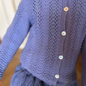 Op de afbeelding: Een periwinkelblauwe gebreide cardigan met een geschulpte vormgeving en drie parelmoer knopen. De trui met lange mouwen heeft een textuur met open weefsel. De cardigan wordt gecombineerd met een bijpassende blauwe rok.