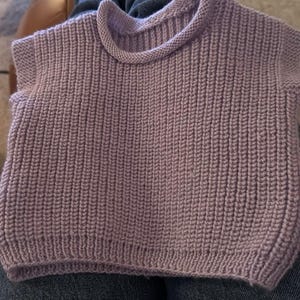 Op de afbeelding: Een lila handgebreide trui met een geribbelde textuur. De trui heeft een ronde hals en korte mouwen. De gebreide stof heeft een verticaal ribpatroon, waardoor een textuur ontstaat. De zoom en de halslijn zijn afgewerkt met een gladde, gerolde rand.