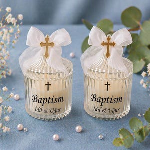 May include: Two glass candle holders with a cream-colored candle inside. Each candle has a gold cross and white ribbon bow. The text "Baptism" and a name are printed on the front. Pearls and white flowers are scattered around.