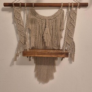 Puede incluir: Una estantería de pared de macramé con una balda de madera y una barra de madera para colgar. La estantería es de macramé beige y tiene una franja decorativa. Se suspende con dos cuerdas.