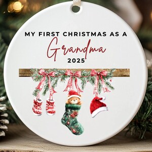 Puede incluir: Adorno navide&ntilde;o de cer&aacute;mica blanca con el texto "MY FIRST CHRISTMAS AS A Grandma 2025". El adorno presenta una viga de madera con decoraciones navide&ntilde;as colgantes, que incluyen mitones, un calcet&iacute;n y un gorro de Pap&aacute; Noel.