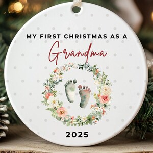 Puede incluir: Adorno navide&ntilde;o redondo de cer&aacute;mica blanca con el texto "MY FIRST CHRISTMAS AS A Grandma". Presenta una corona floral con huellas de beb&eacute; y el a&ntilde;o 2025.