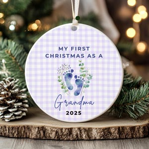 Puede incluir: Adorno navide&ntilde;o redondo de cer&aacute;mica con un patr&oacute;n de cuadros vichy en lila claro. El adorno tiene el texto "MY FIRST CHRISTMAS AS A Grandma 2025" y presenta huellas de pies azules con detalles florales. Una cinta blanca est&aacute; adjunta para colgar.