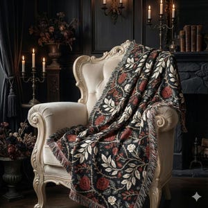 May include: A decorative throw blanket draped over a cream-colored armchair. The blanket features a dark background with a repeating pattern of red roses, white leaves, and vines. The edges of the blanket have a fringed trim. The room has a dark, gothic aesthetic.
