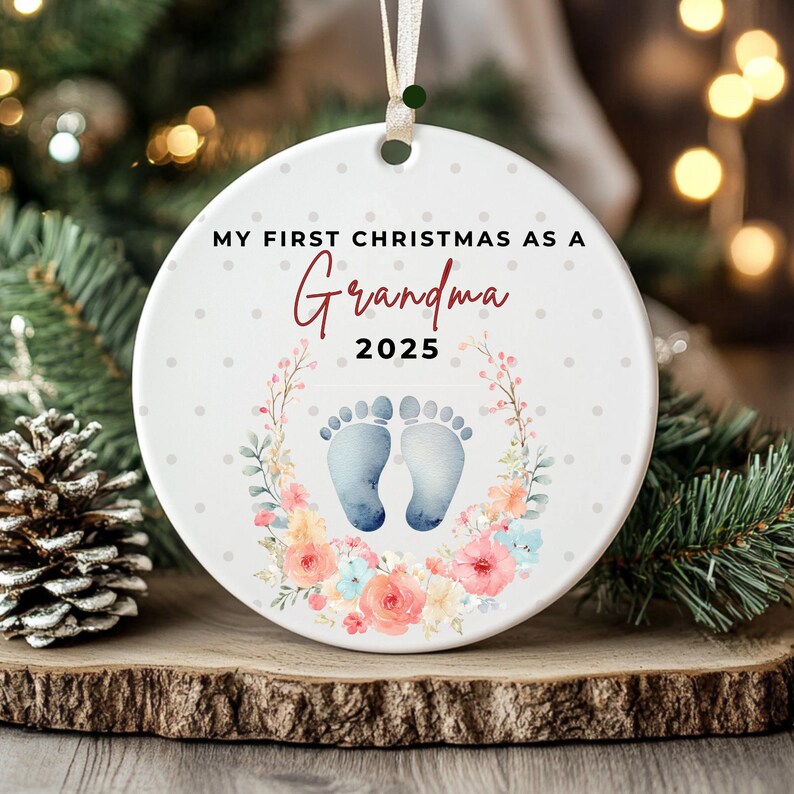 Puede incluir: Adorno navide&ntilde;o redondo de cer&aacute;mica blanca con el texto "MY FIRST CHRISTMAS AS A Grandma 2025". Presenta huellas de beb&eacute; y una corona floral. Cuelga de una cinta blanca.