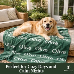 以下が含まれることがあります： 白い文字で「Olive」と繰り返された緑色の毛布の上に、ゴールデンレトリバー犬がいます。毛布は籐の椅子に掛けられています。「Perfect for Cozy Days and Calm Nights」というテキストが下にあります。
