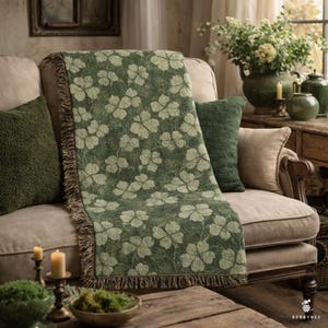 May include: A green clover-patterned throw blanket with a fringed edge, draped over a beige sofa. The scene includes green and beige cushions, candles, and floral arrangements, creating a cosy living room setting.